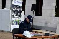 Fotografia kolorowa przedstawiająca Komendanta Miejskiego  Policji w Białymstoku insp. Wojciecha Macutkiewicza   podpisującego akt erekcyjny przy stoliku.
