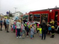 Fotografia kolorowa przedstawiająca przedszkolaki stojące przy samochodzie bojowym Państwowej Straży Pożarnej, którym prezentowany jest sprzęt pożarniczy.