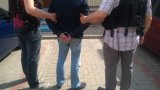 Fotografia kolorowa przedstawiająca zatrzymanego z kajdankami założonymi na ręce trzymane z tyłu prowadzonego przez dwóch policjantów