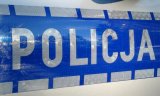 Fotografia kolorowa przedstawiająca srebrny napis policja na niebieskim tle. Umiejscowiony na bocznych drzwiach radiowozu