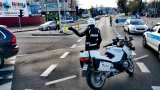 Fotografia kolorowa przedstawiająca na pierwszym planie policjanta ruchu drogowego stojącego przy motocyklu policyjnym kierujący ruchem. W tle inni policjanci stojący w rejonie skrzyżowania