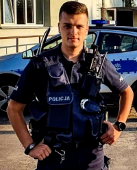 Fotografia kolorowa przedstawiająca policjanta stojącego na tle radiowozu