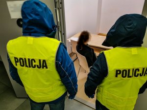 Policjanci z wydziału kryminalnego Komisariatu Policji III w Białymstoku ubrani w żółte kamizelki z napisem na plecach: &quot; policja&quot;. Policjanci stoją przed pomieszczeniem, w którym na krześle siedzi zatrzymana kobieta.