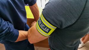 policjant w cywilnych ubraniach z żółtą opaską z napisem&quot;policja&quot;, zakłada mężczyźnie kajdanki na ręce trzymane z tyłu