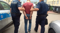 Fotografia kolorowa przedstawiająca zatrzymanego z kajdankami na rękach trzymanych z tyłu, prowadzonego przez policjantów