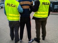 Fotografia kolorowa przedstawiająca zatrzymanego w kajdankach zespolonych doprowadzanego przez policjantów.