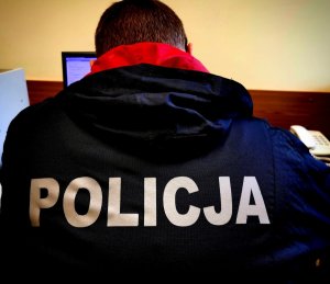 policjant siedzący plecami do zdjęcia przy biurku. Policjant ubrany w czarna kurtkę z napisem &quot;policja&quot;