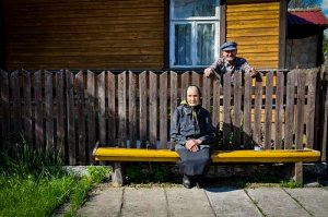 starsza kobieta siedząca na drewnianej ławce przed domem. Za nią za płotem stoi starszy mężczyzna