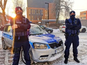 Umundurowani policjanci stojący przy radiowozie, w tle zimowa aura.