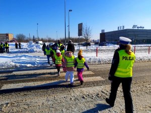 Grupa dzieci przechodząca w kamizelkach odblaskowych przez przejście dla pieszych, na końcu idąca policjantka