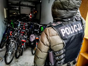 policjant w cywilnych ubraniach, z założoną czarną kamizelką z napisem &quot;POLICJA&quot;. Policjant stoi przy garażu, w którym stoją odzyskane rowery