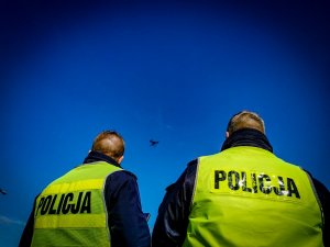 dwóch policjantów w żółtych, odblaskowych kamizelkach, którzy obserwują latającego drona