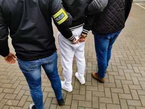 Policjanci kryminalni w ubraniach cywilnych trzymają zatrzymaną osobę, która ma założone kajdanki na ręce trzymane z tyłu
