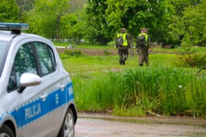 Funkcjonariusze Wojska Obrony Terytorialnej przeczesujący tereny zielone w poszukiwaniu zaginionej kobiety, w tle radiowóz policyjny