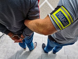 policjant kryminalny w cywilnych ubraniach  z założona opaską z napisem &quot;POLICJA&quot;. Policjant trzyma zatrzymanego, który ma założone kajdanki na rękach trzymanych z tyłu.