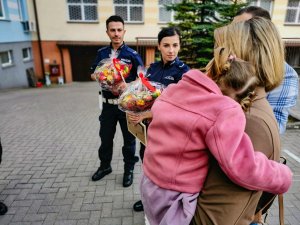 patrol policjantów z białostockiej drogówki, który pilotował rodziców z dzieckiem do szpitala. Policjanci trzymają ramkę z rysunkiem oraz bukiety warzywno-owocowe . Na pierwszym planie mama trzymająca na rękach dziewczynkę