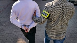 policjant kryminalny z założoną opaską z napisem &quot;POLICJA&quot;. Funkcjonariusz prowadzi zatrzymanego mężczyznę
