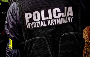 Kryminalny w cywilnych ubraniach z założoną czarna kamizelką z napisem: POLICJA WYDZIAŁ KRYMINALNY&quot;