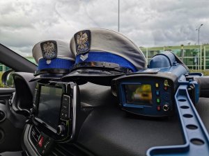 dwie policyjne czapki z Wydziału Ruchu Drogowego, obok urządzenie do pomiaru prędkości. Rzeczy ułożone w środku radiowozu.
