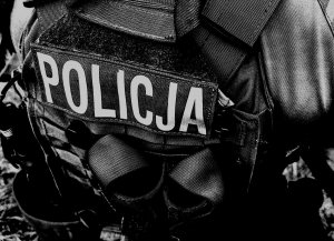 zdjęcie wykonane w szarości, które przedstawia plecy policjant a z kamizelką taktyczną z napisem &quot;POLICJA&quot;