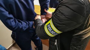 policjantka w cywilnych ubraniach z założoną żółtą opaską z napisem &quot;POLICJA&quot;. Policjantka zakłada mężczyźnie kajdanki na ręce trzymane z tyłu