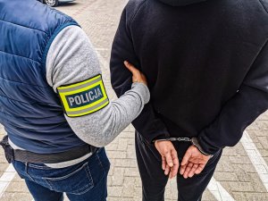 Nieumundurowany policjant z opaską na ramieniu z napisem Policja, prowadzi zatrzymanego mężczyznę.