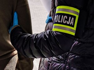 Policjant z opaską na ramieniu z napisem POLICJA trzymający zatrzymanego