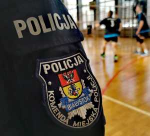 Rękaw munduru z logo Komendy Miejskiej Policji w Białymstoku, w tle rozgrywany mecz siatkówki.