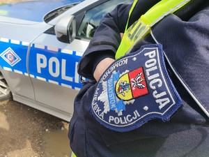 Rękaw kurtki z tarczą policyjną, w tle radiowóz policyjny.