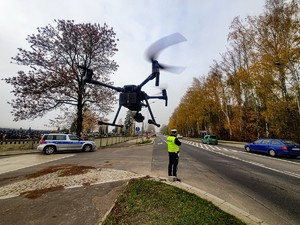 Policjant mierzący prędkość ręcznym miernikiem prędkości, nad nim zawieszony w powietrzu dron.