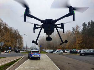 Dron zawieszony w powietrzu, w tle radiowóz.