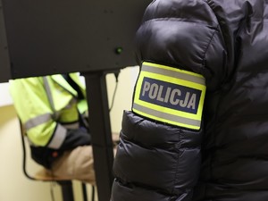 Ręka policjanta z opaską z napisem policja, w tle zatrzymany siedzący na krześle.