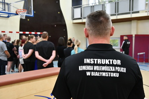 Mężczyzna stojący tyłem z napisem instruktor Komenda Wojewódzka Policji w Białymstoku, w tle inne osoby.