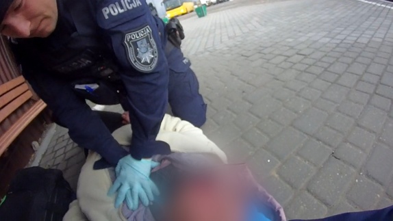 Zdjęcie przedstawia policjanta, który prowadzi resuscytacje kobiecie leżącej na przystanku autobusowym.